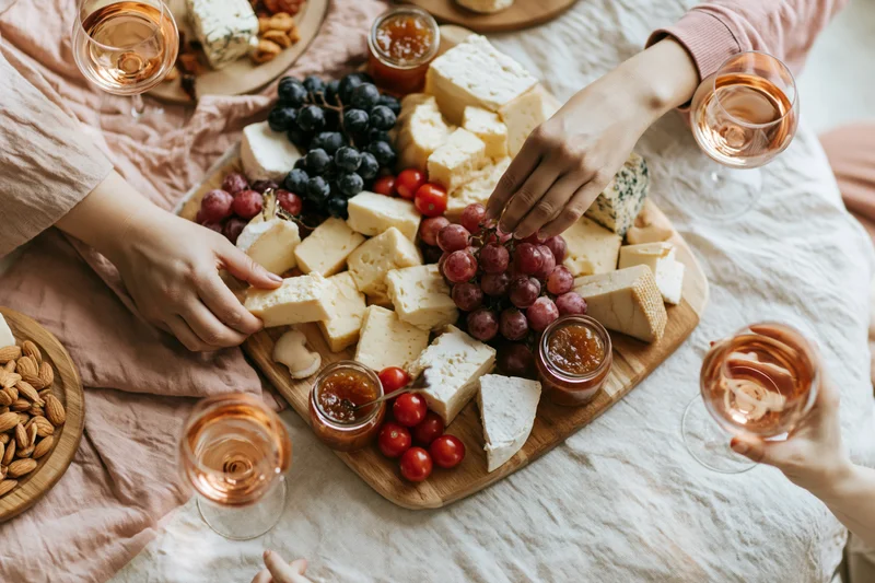 Hands reaching across a charcuterie board with cheese, grapes, and wine on a tablecloth