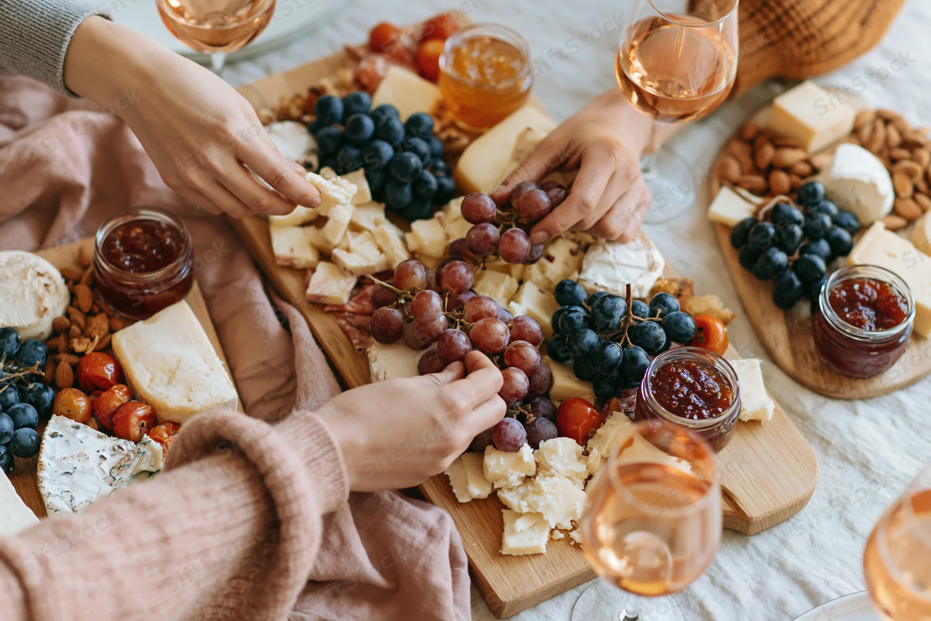 Hands reaching across a charcuterie board with cheese, grapes, and wine on a table