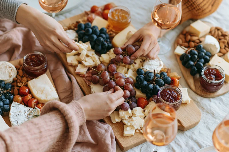 Hands reaching across a charcuterie board with cheese, grapes, and wine on a table
