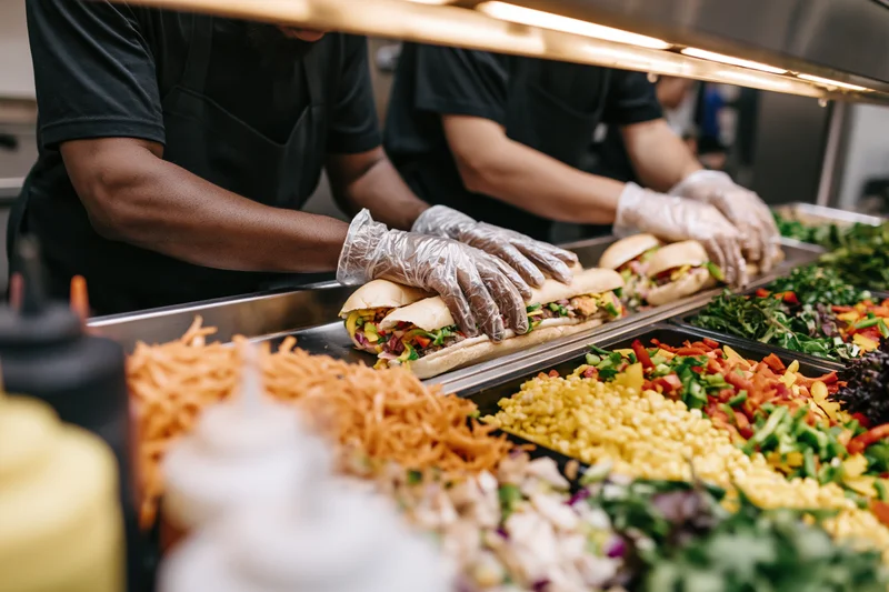Workers assembling sandwiches with fresh vegetables and meats at a deli counter