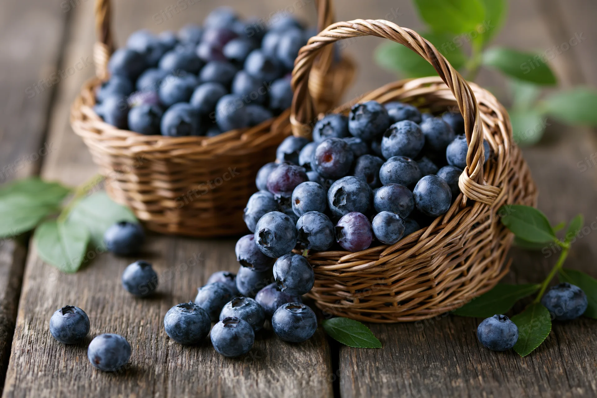 Two wicker baskets filled with fresh blueberries on a wooden table with scattered berries and green leaves