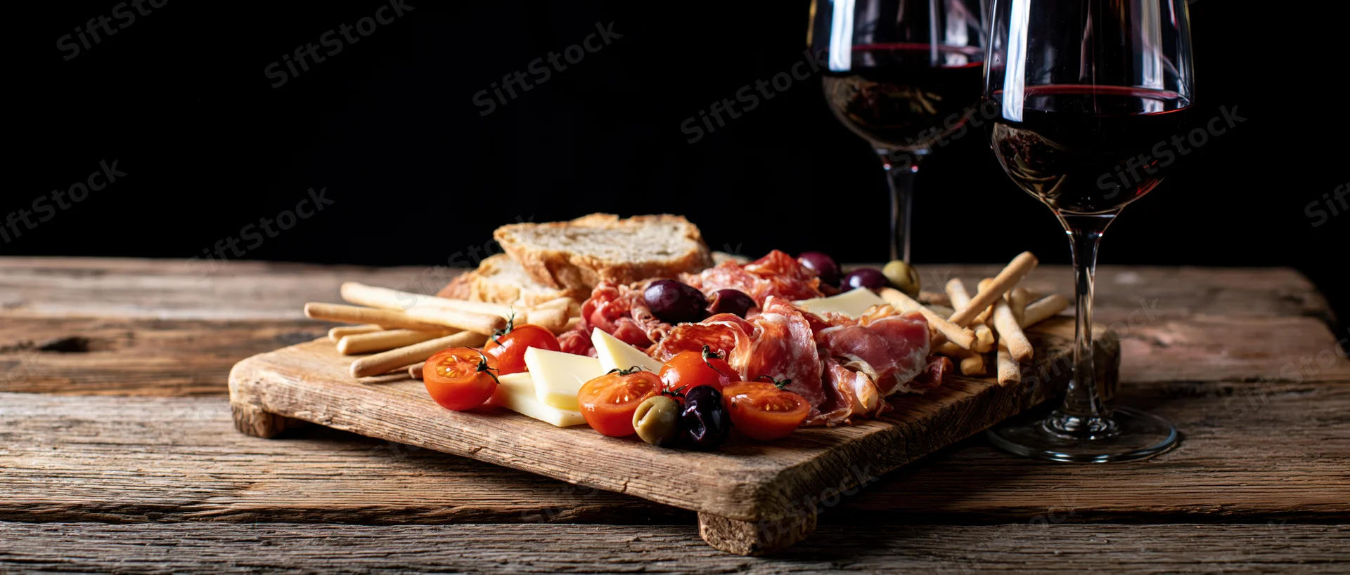 Two stemmed glasses of red wine beside a rustic wooden board with cheese, cured meats, tomatoes, olives, bread and crackers on a dark wooden table