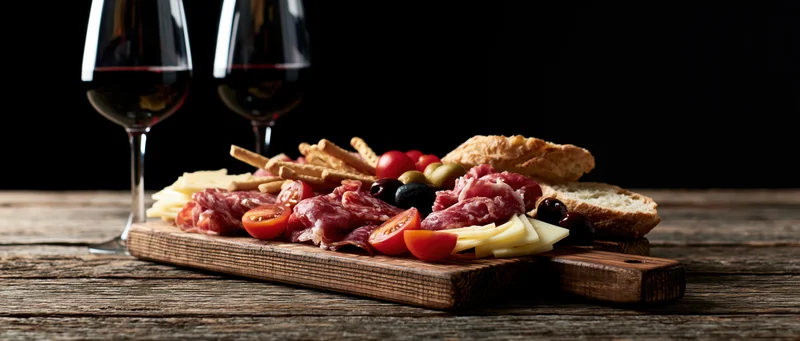 Charcuterie board with cheese, cured meats, tomatoes, olives, bread and crackers beside two glasses of red wine on a rustic wooden table