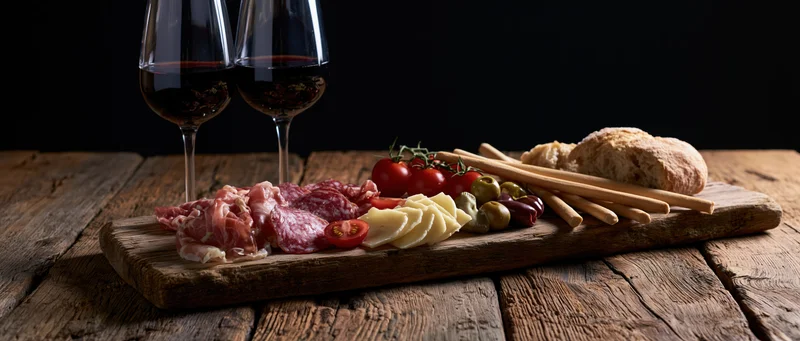 Two stemmed glasses of red wine beside a rustic wooden board with cheese, cured meats, tomatoes, olives, bread and crackers