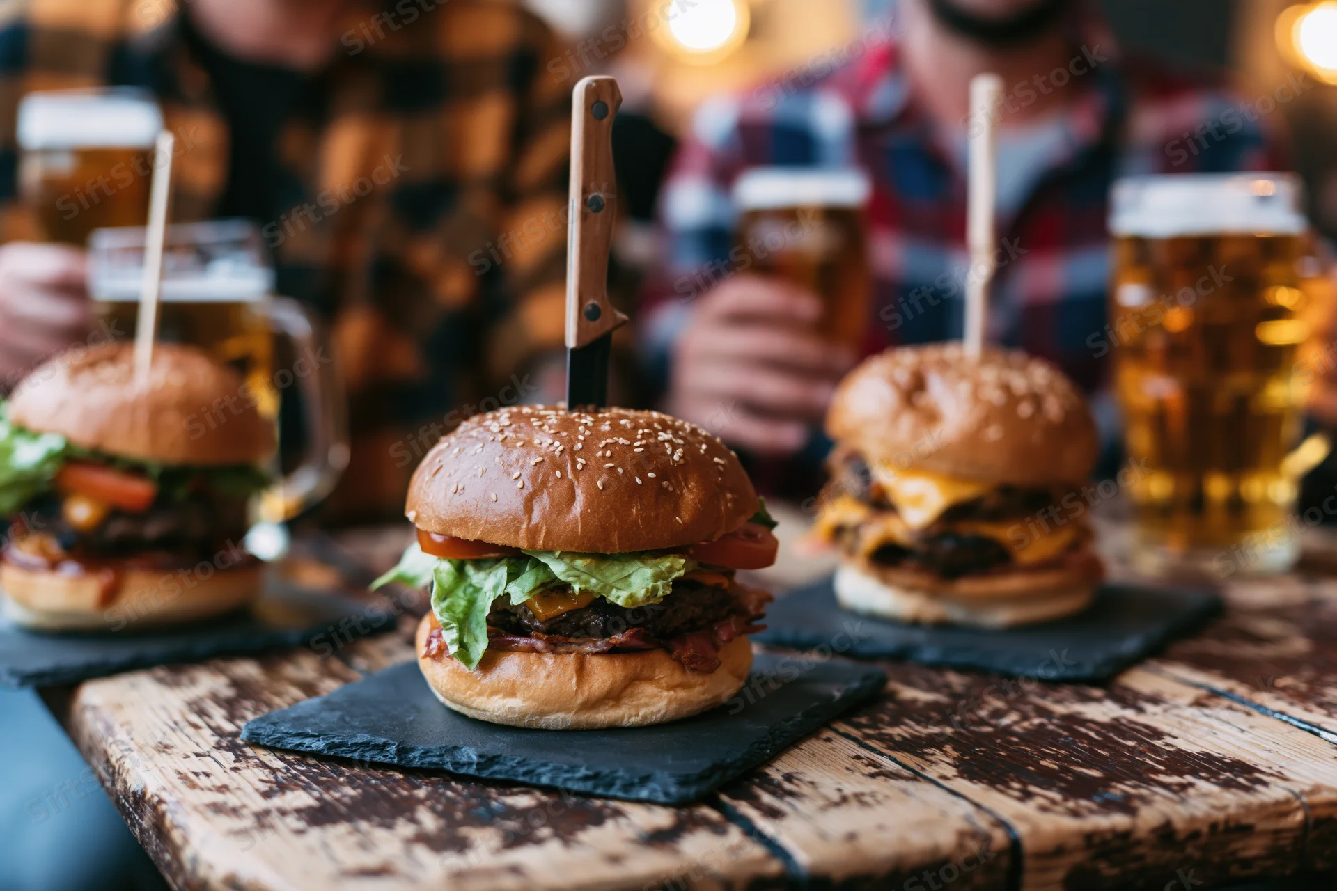 Three stacked burgers on slate platters with beer mugs on a wooden table, served at a casual dining setting