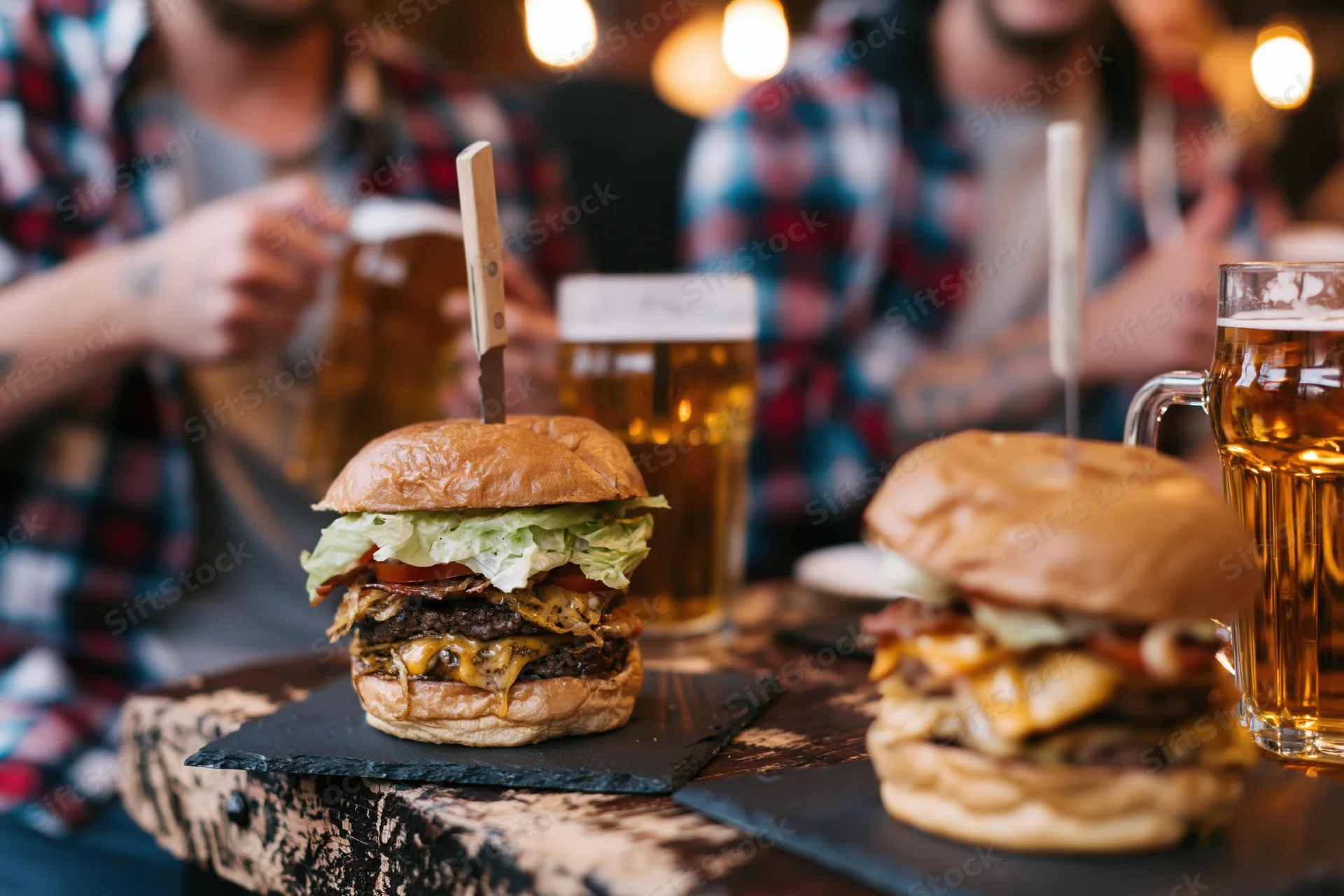 Two tall stacked burgers on brioche buns served on slate plates with beer glasses on a wooden table in a dimly lit pub setting