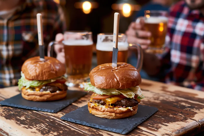 Two cheeseburgers on brioche buns with knives inserted, served on slate platters on a wooden table with beer mugs in the background