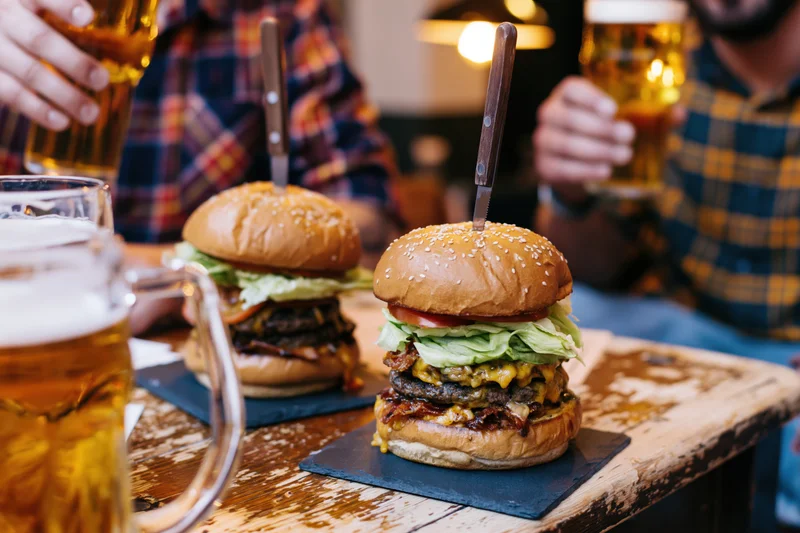 Two stacked burgers with bacon and cheese on brioche buns served on slate plates with beer mugs on a wooden table