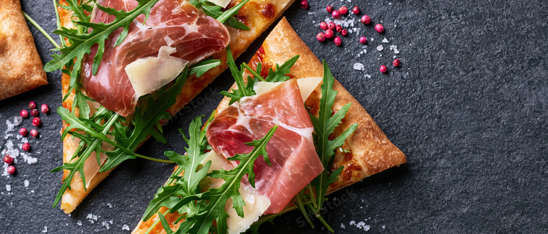 Two triangular pizza slices topped with prosciutto and arugula on a dark slate surface