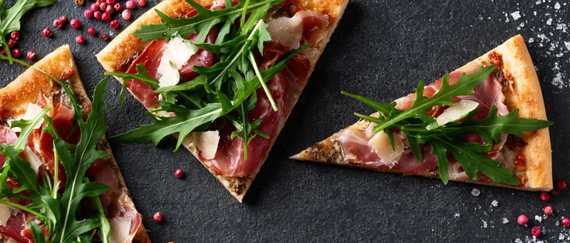 Three triangular pizza slices topped with prosciutto and arugula on a dark slate surface