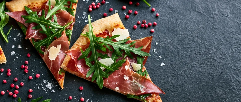 Triangular pizza slices topped with prosciutto and arugula on a dark slate surface