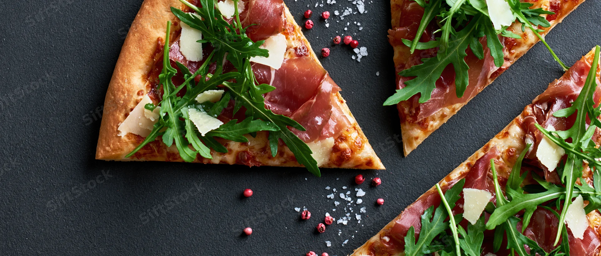 Triangular pizza slices topped with prosciutto and arugula on a black slate surface