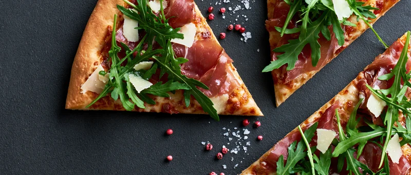 Triangular pizza slices topped with prosciutto and arugula on a black slate surface