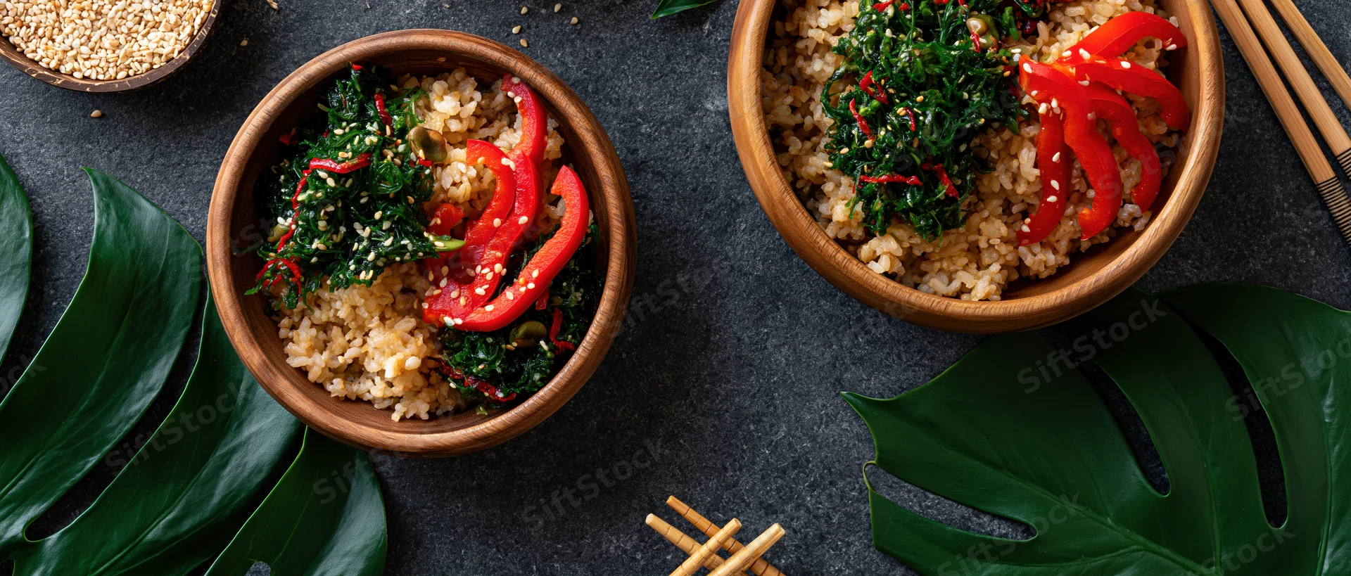 Two wooden bowls filled with brown rice and red bell pepper slices on dark surface with monstera leaves and chopsticks