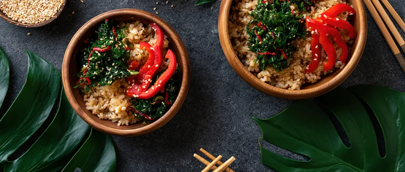Two wooden bowls filled with brown rice and red bell pepper slices on dark surface with monstera leaves and chopsticks