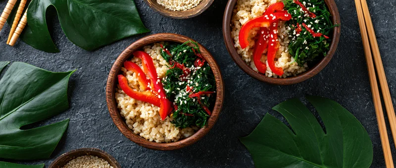 Two wooden bowls filled with brown rice, red bell pepper slices, spinach, and sesame seeds on a dark surface with monstera leaves and chopsticks