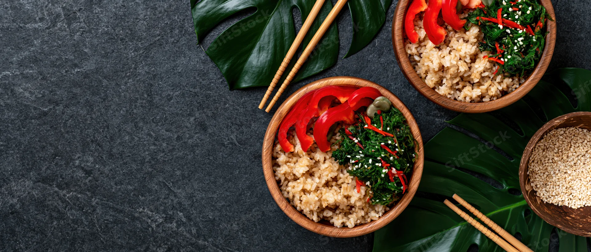 Wooden bowls with brown rice and red bell pepper slices arranged with chopsticks and monstera leaves on dark stone surface