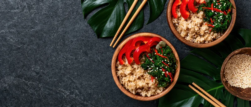 Wooden bowls with brown rice and red bell pepper slices arranged with chopsticks and monstera leaves on dark stone surface