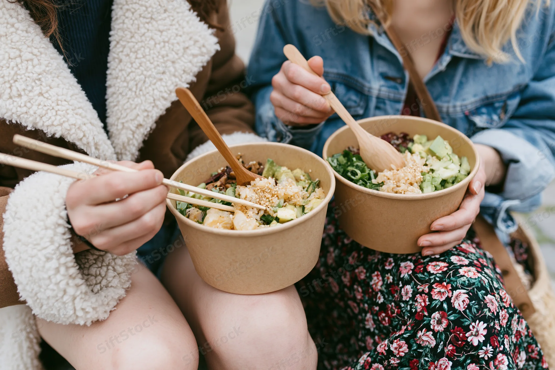 Two young women eating poke bowls outdoors while sitting together