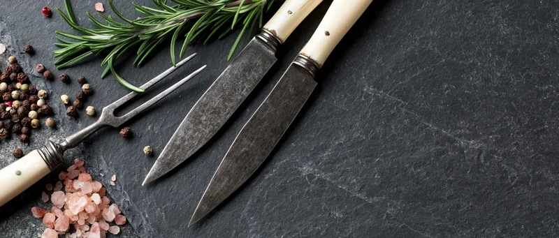 Vintage knife and fork with bone-white handles resting on dark slate surface beside rosemary, peppercorns and pink salt