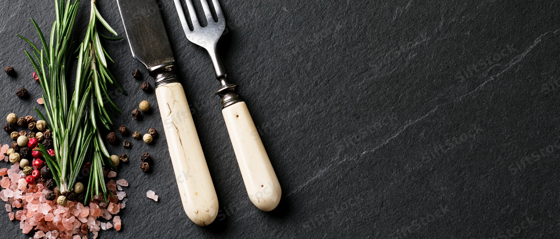 Vintage knife and fork with bone-white handles resting beside rosemary, peppercorns and pink salt on dark slate surface