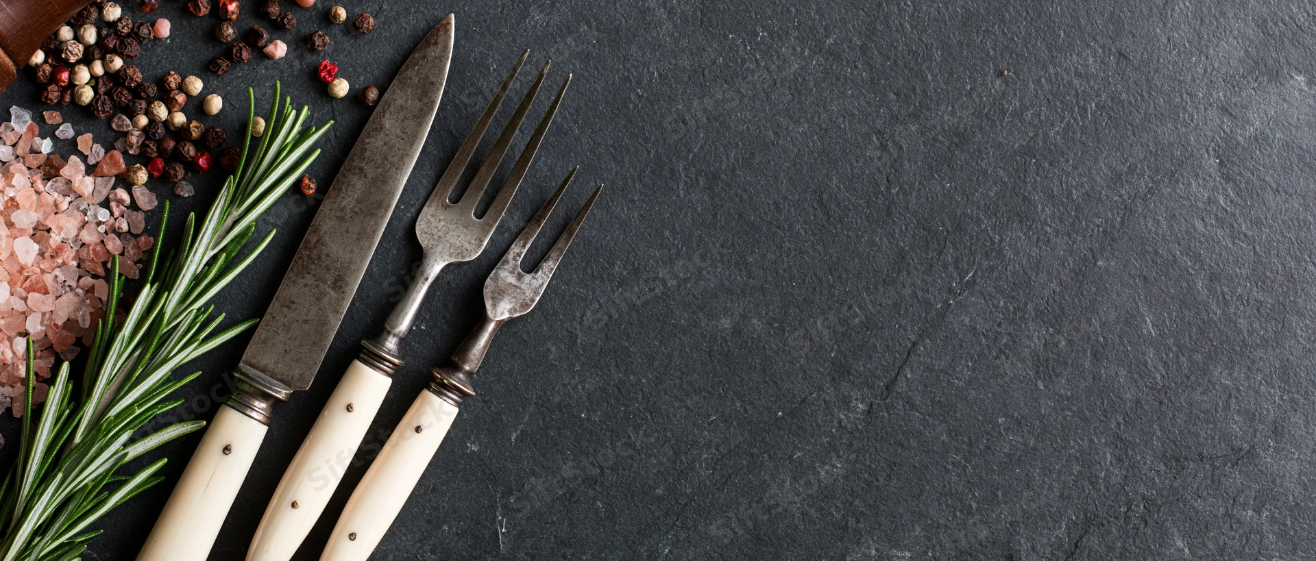 Vintage knife and fork with bone handles arranged with herbs and spices on dark slate surface