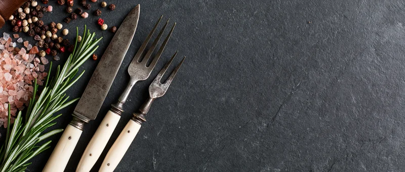 Vintage knife and fork with bone handles arranged with herbs and spices on dark slate surface
