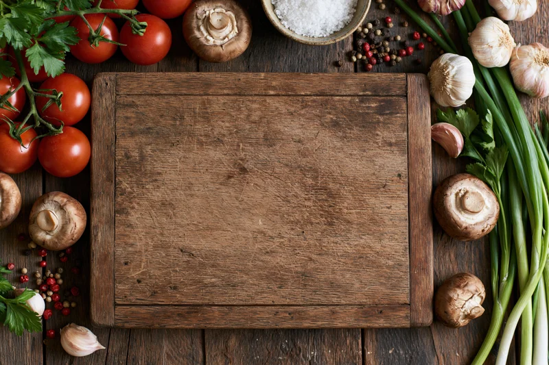 Weathered wooden cutting board surrounded by fresh vegetables and herbs on dark rustic countertop