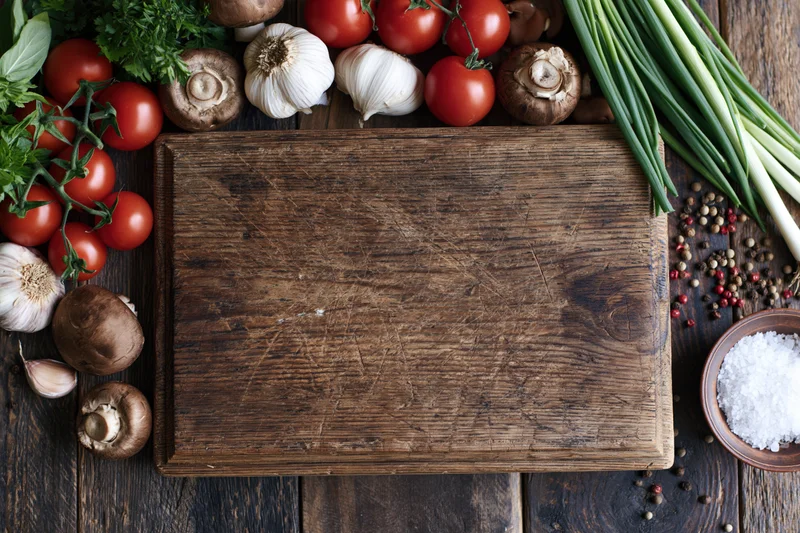 Weathered wooden cutting board surrounded by fresh vegetables and herbs on dark rustic table