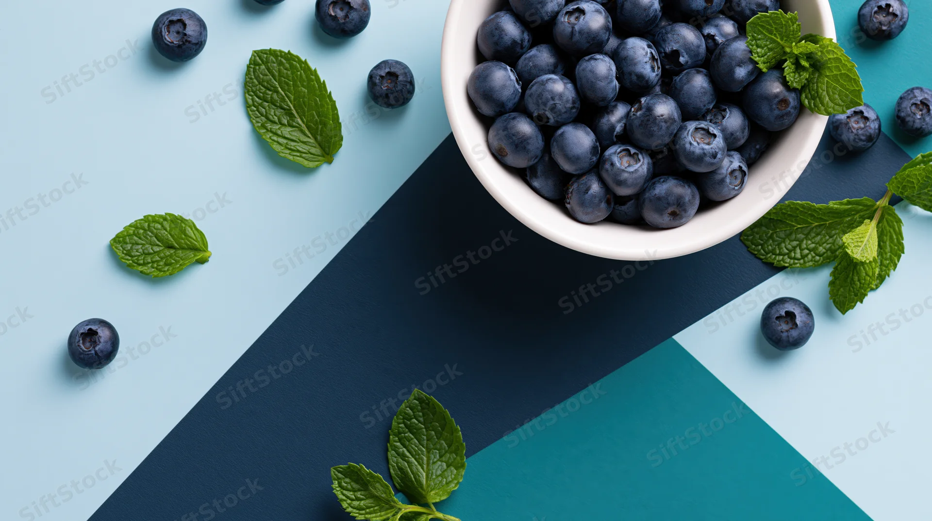 White ceramic bowl filled with fresh blueberries and mint leaves on a geometric blue surface, top view flat lay arrangement