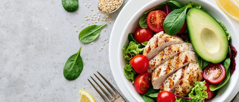 Grilled chicken and avocado salad in a white bowl with fresh vegetables and lemon on a gray surface