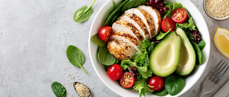Grilled chicken and avocado salad in a white bowl with spinach and cherry tomatoes on a light surface