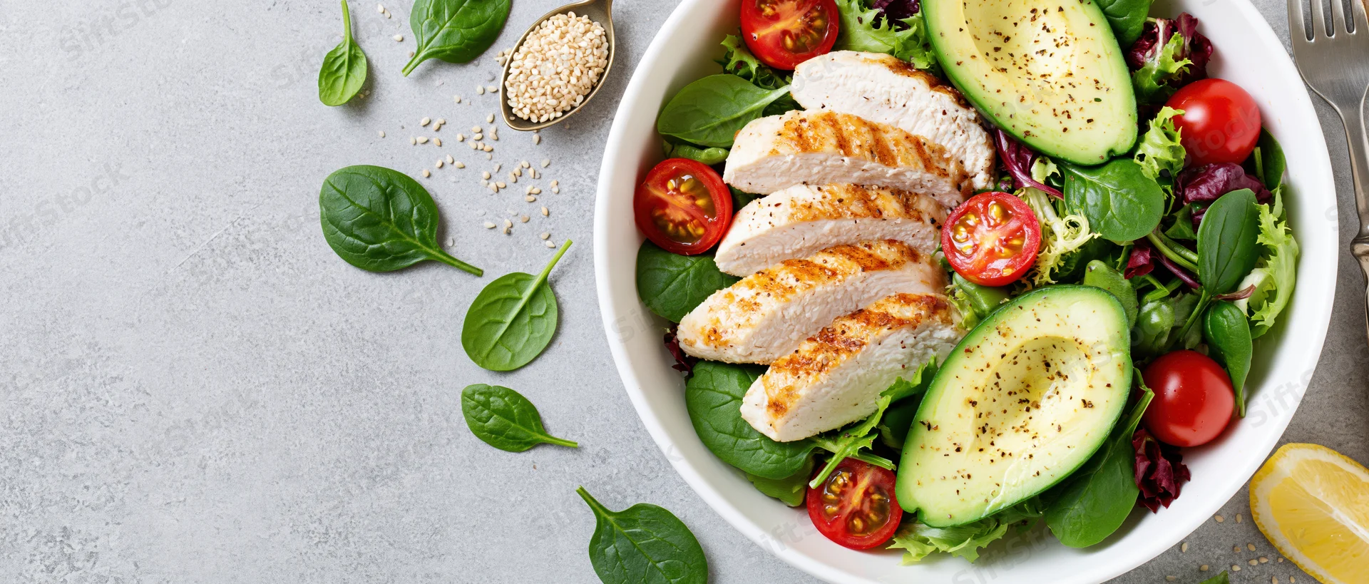 Grilled chicken and avocado salad in a white bowl with spinach and tomatoes on a gray surface