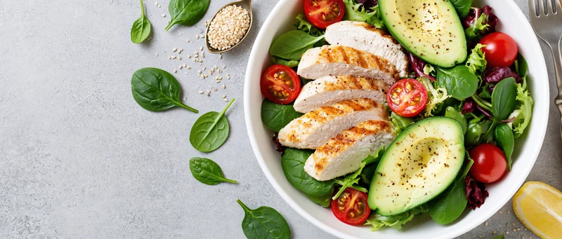 Grilled chicken and avocado salad in a white bowl with spinach and tomatoes on a gray surface