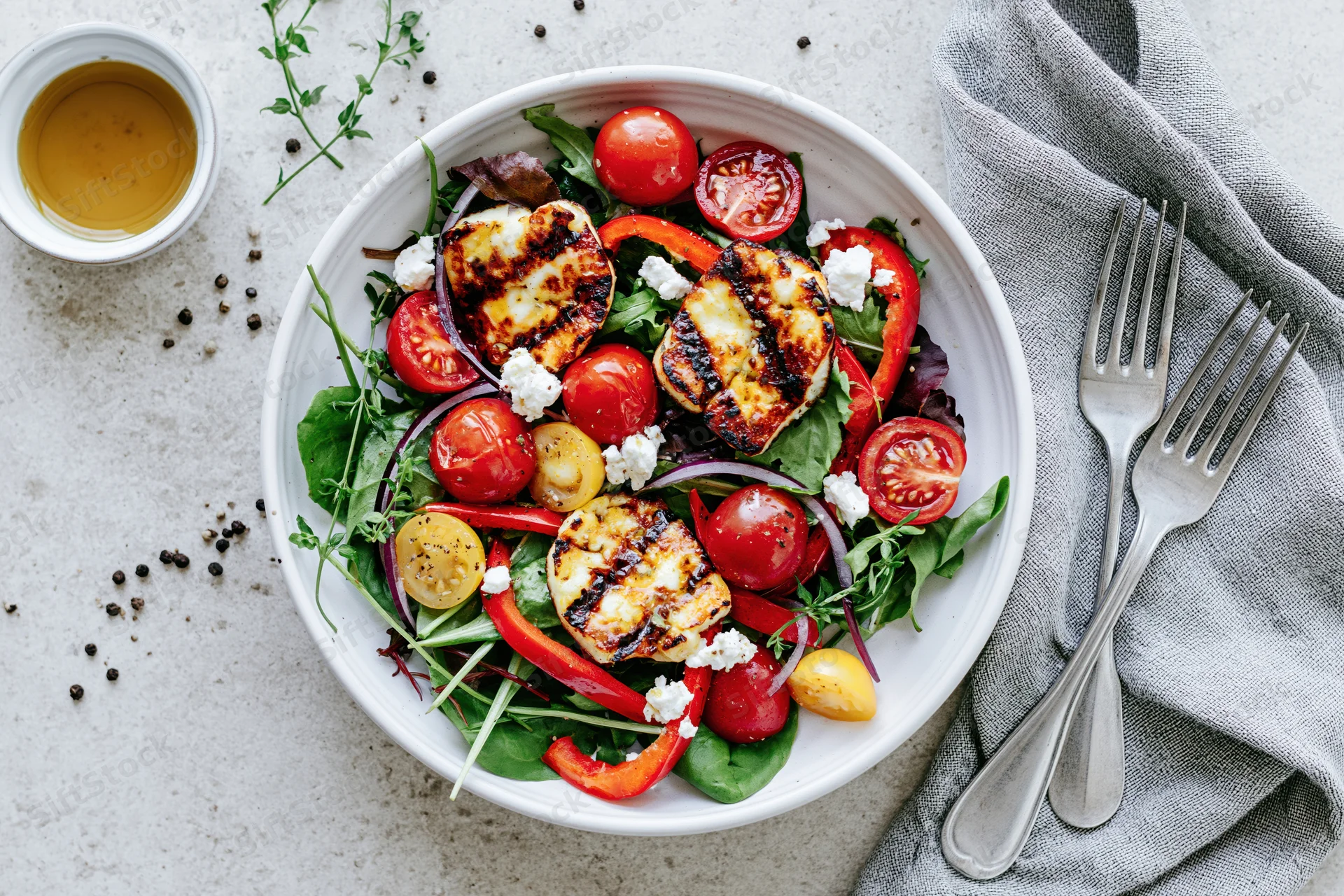 Grilled halloumi salad with cherry tomatoes and peppers in a white ceramic bowl on a countertop with cutlery and napkin