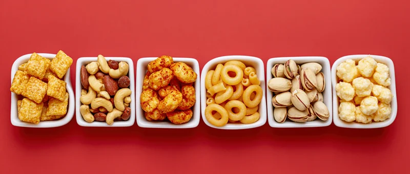 Six white ceramic dishes arranged in a row on red background each filled with different snack foods