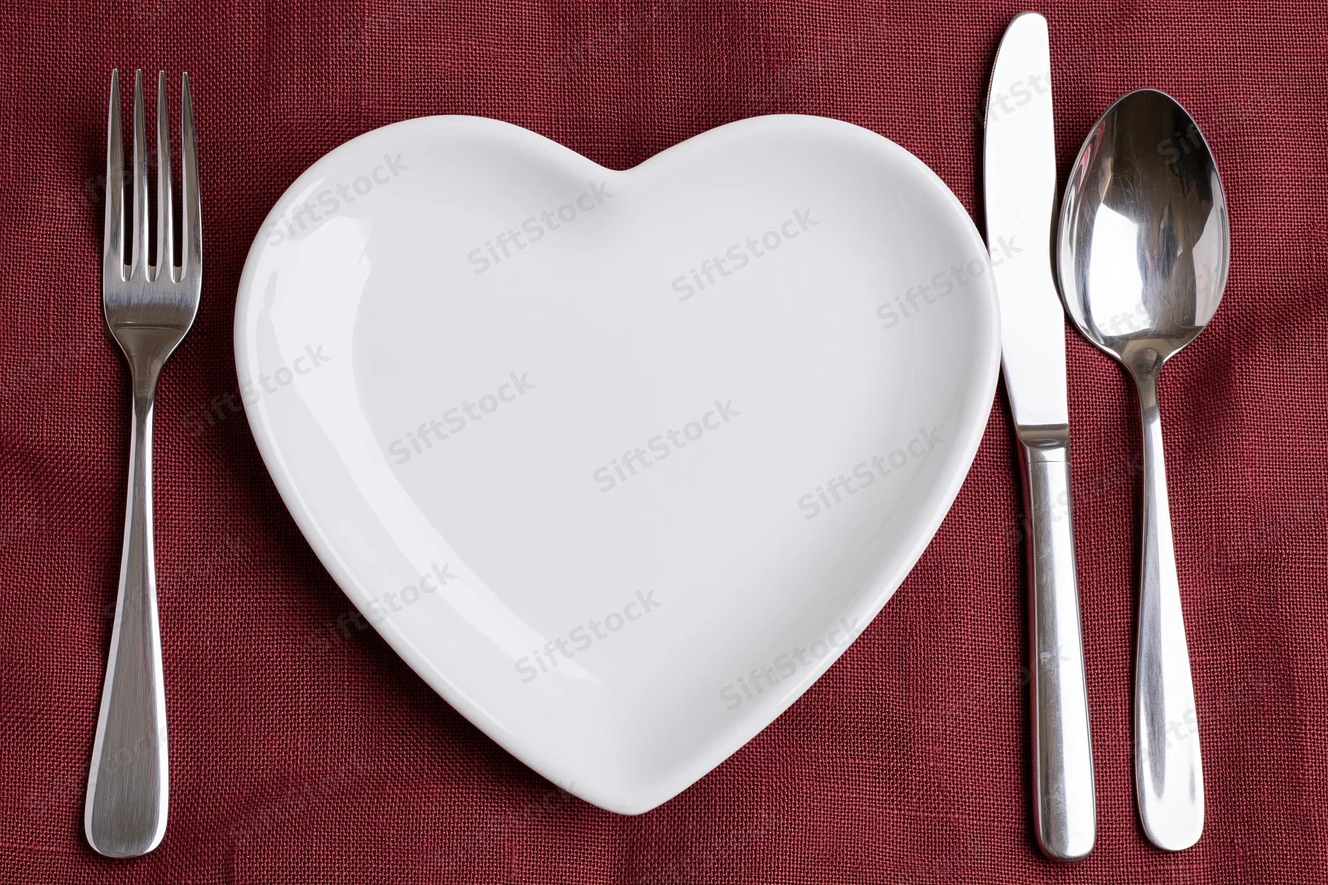 Heart-shaped white ceramic plate centered on red linen tablecloth with fork, knife and spoon arranged around it
