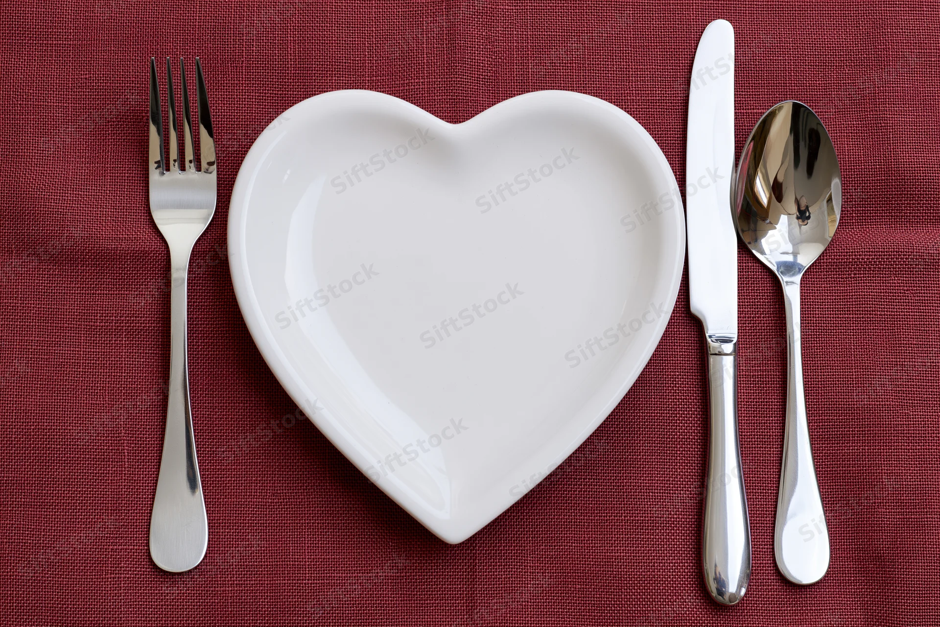 Heart-shaped white ceramic plate with silverware arranged on red linen tablecloth