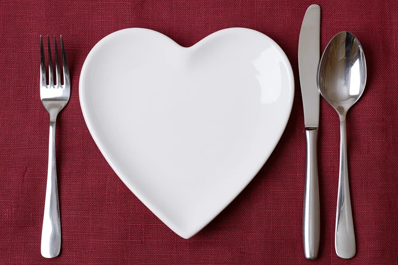 Heart-shaped white ceramic plate centered on red linen tablecloth with fork, knife and spoon arranged around it