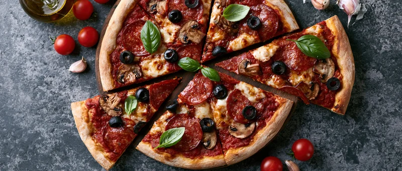 Sliced pepperoni and mushroom pizza on dark stone surface with cherry tomatoes and garlic cloves scattered around