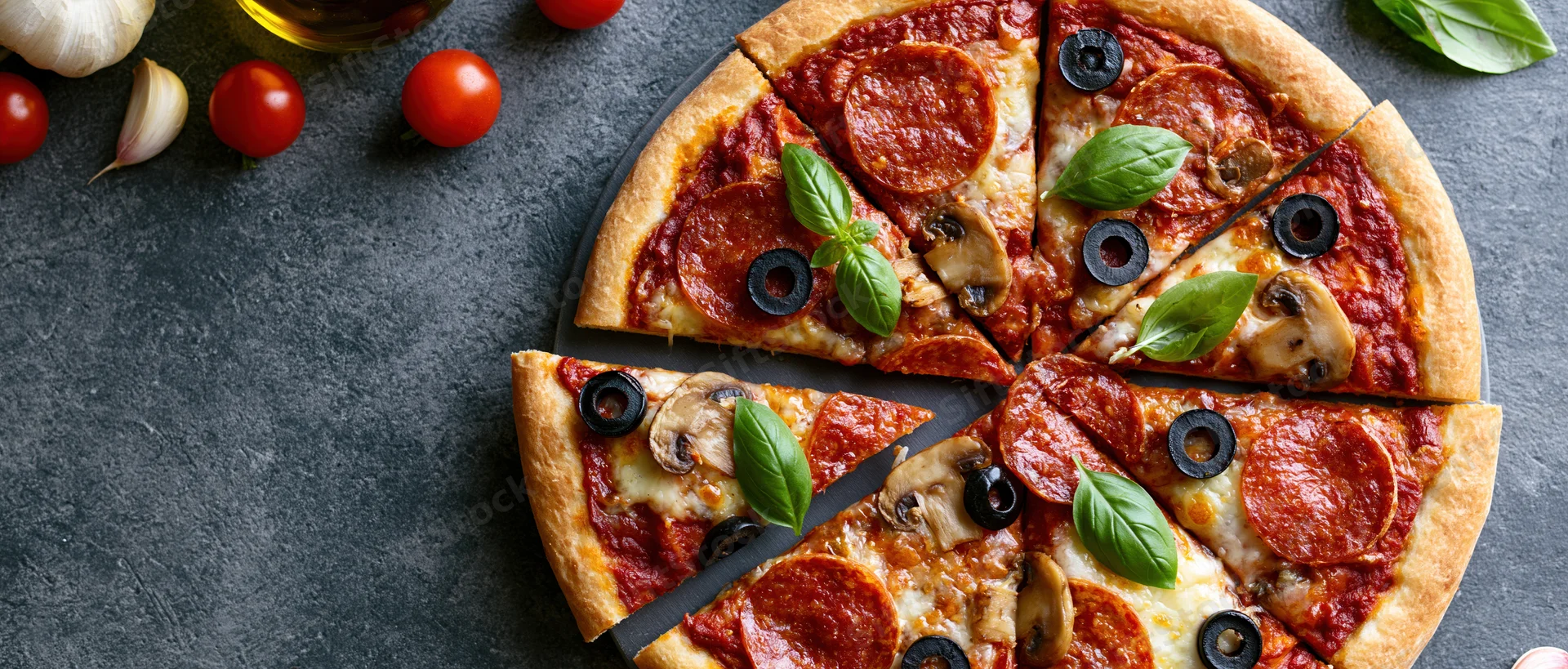 Sliced pepperoni and mushroom pizza on dark stone surface with fresh ingredients arranged around