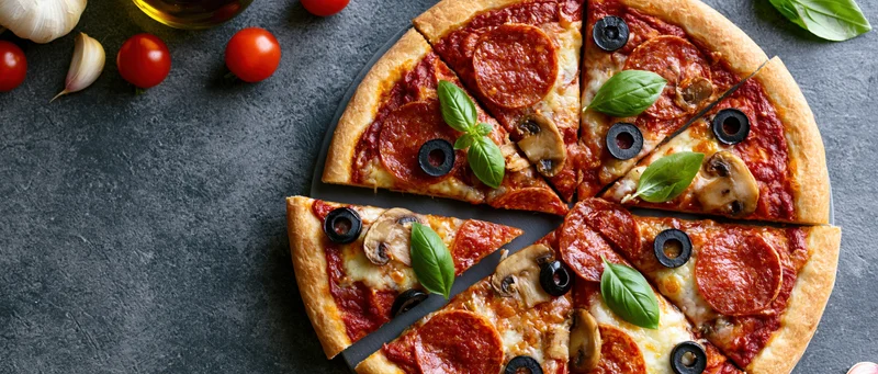 Sliced pepperoni and mushroom pizza on dark stone surface with fresh ingredients arranged around