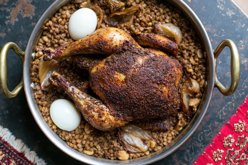 Whole roasted spiced chicken in a brass-handled pot with lentils and eggs on a patterned textile surface