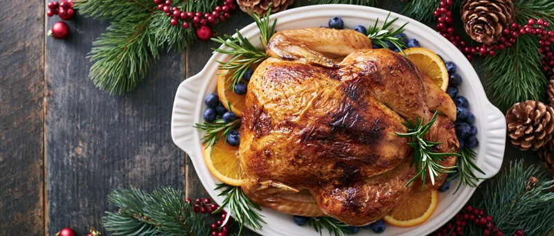 Roasted turkey on a white ceramic platter garnished with orange slices, rosemary, and blueberries, surrounded by pine branches and red berries on a dark wooden table