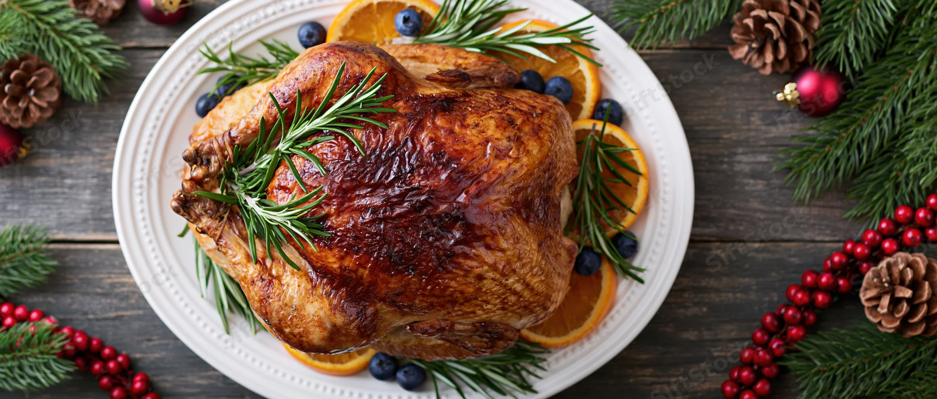Roasted turkey on a white platter garnished with rosemary, orange slices and blueberries surrounded by christmas decorations on a wooden table