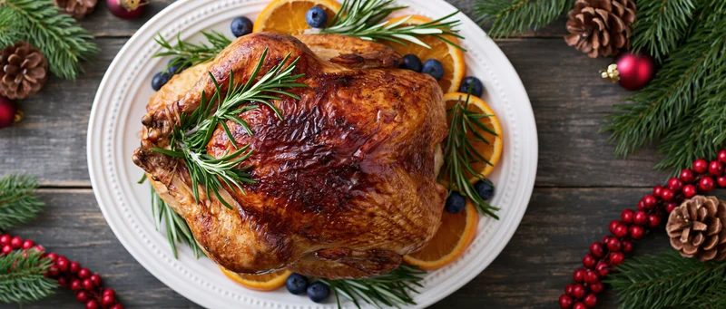 Roasted turkey on a white platter garnished with rosemary, orange slices and blueberries surrounded by christmas decorations on a wooden table