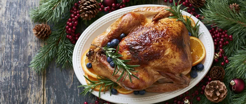 Roasted turkey on a white ceramic platter garnished with rosemary, orange slices and blueberries, surrounded by pine branches and holiday decorations on a wooden table