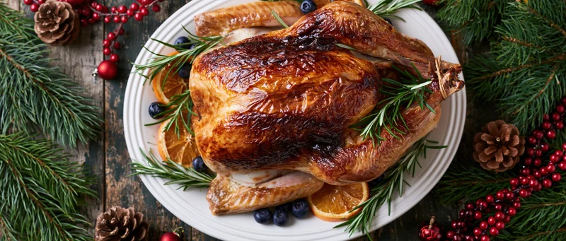 Roasted turkey on a white ceramic platter garnished with rosemary, orange slices, and blueberries surrounded by pine branches and holiday decorations on a rustic wooden table