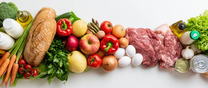 Wide flat-lay arrangement of mixed groceries piled in a panoramic row on white surface