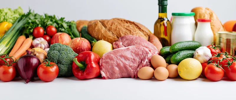 Wide flat-lay arrangement of mixed groceries piled on white surface